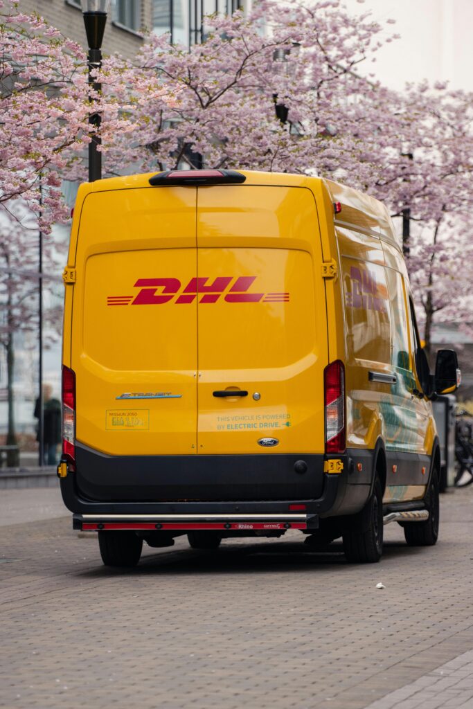 Gele DHL-bezorgbus rijdt door een stedelijke straat met bloeiende bomen, symbool voor duurzame en elektrische pakketbezorging.